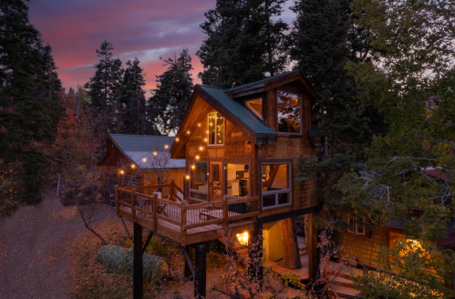 A cozy wooden cabin nestled among trees, illuminated by warm lights at sunset.